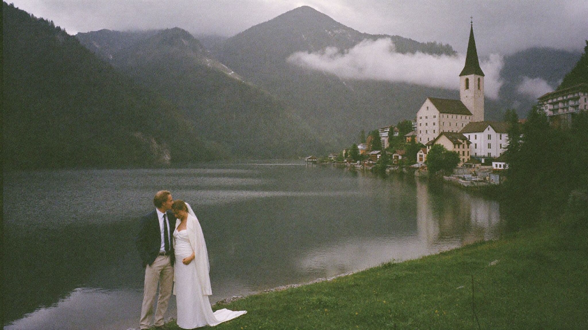 Couple on the lakeshore promenade of Hallstatt at dawn during their Austria elopement, village and mountains reflected in the water