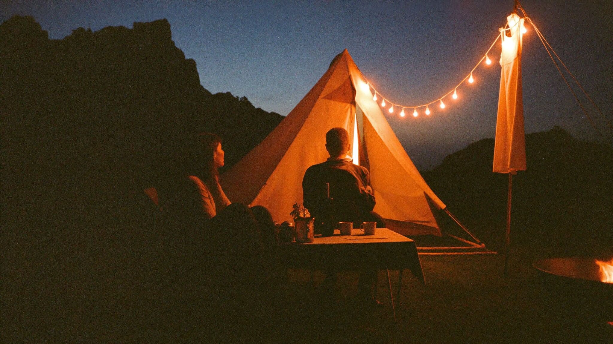 Couple relaxing outside a canvas tent with lanterns at dusk after their elopement