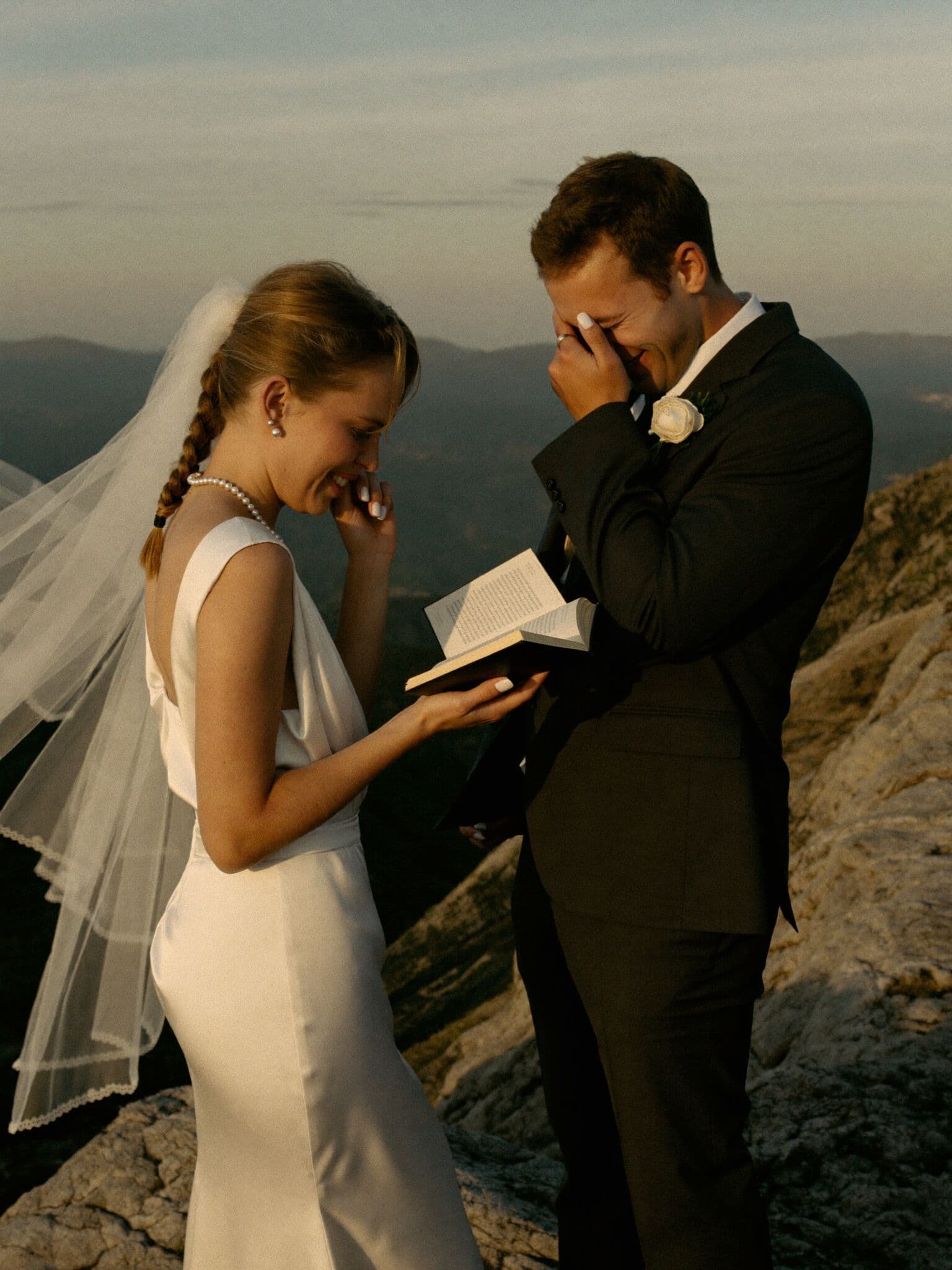 Bride reading handwritten vows on a mountain summit while her groom laughs